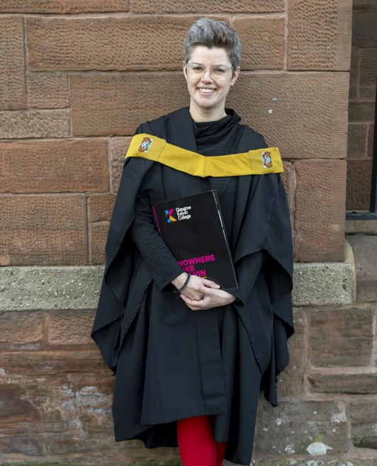 Graduate in gown and gold sash smiling while holding a certificate outside a red-brick building. Graduate in gown and gold sash smiling while holding a certificate outside a red-brick building.